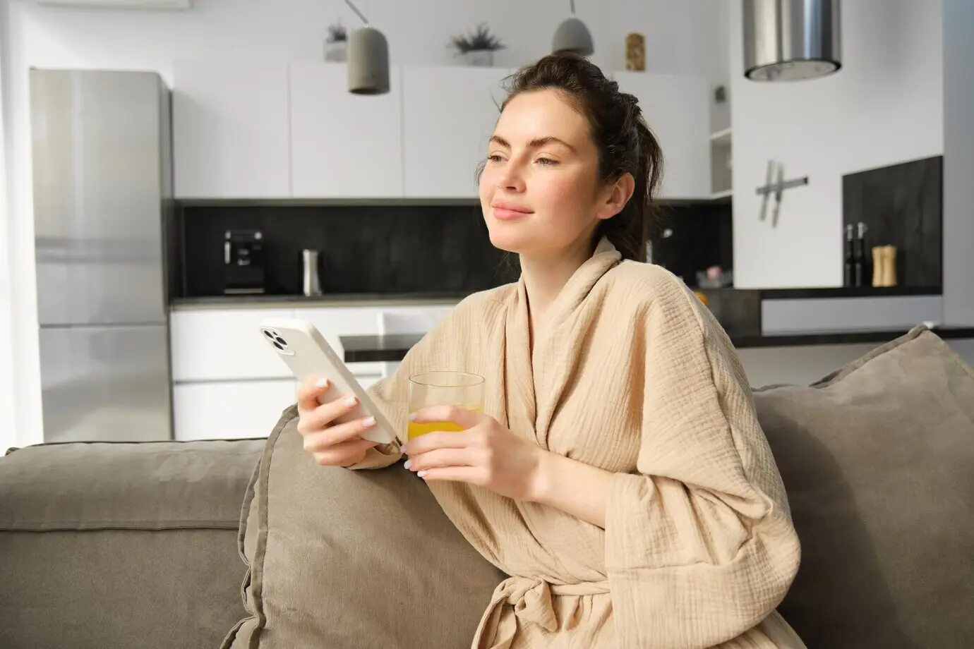 Hermosa mujer joven descansando en el sofá, disfrutando de su mañana en casa, sosteniendo un vaso de jugo de naranja y