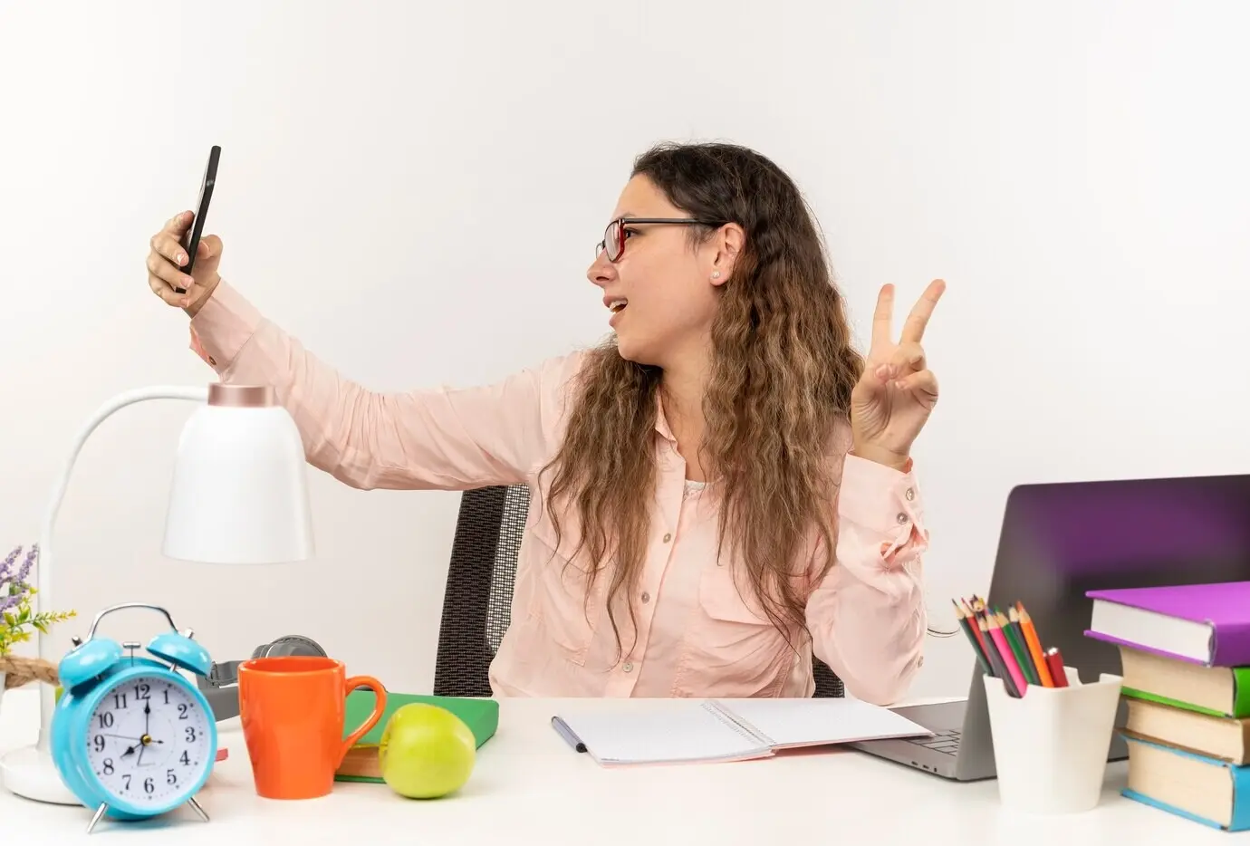 Alegre niña escolar joven y bonita con gafas, sentada en un escritorio con útiles escolares, haciendo su tarea, haciendo el signo de paz y tomándose una selfie, aislada sobre fondo blanco.